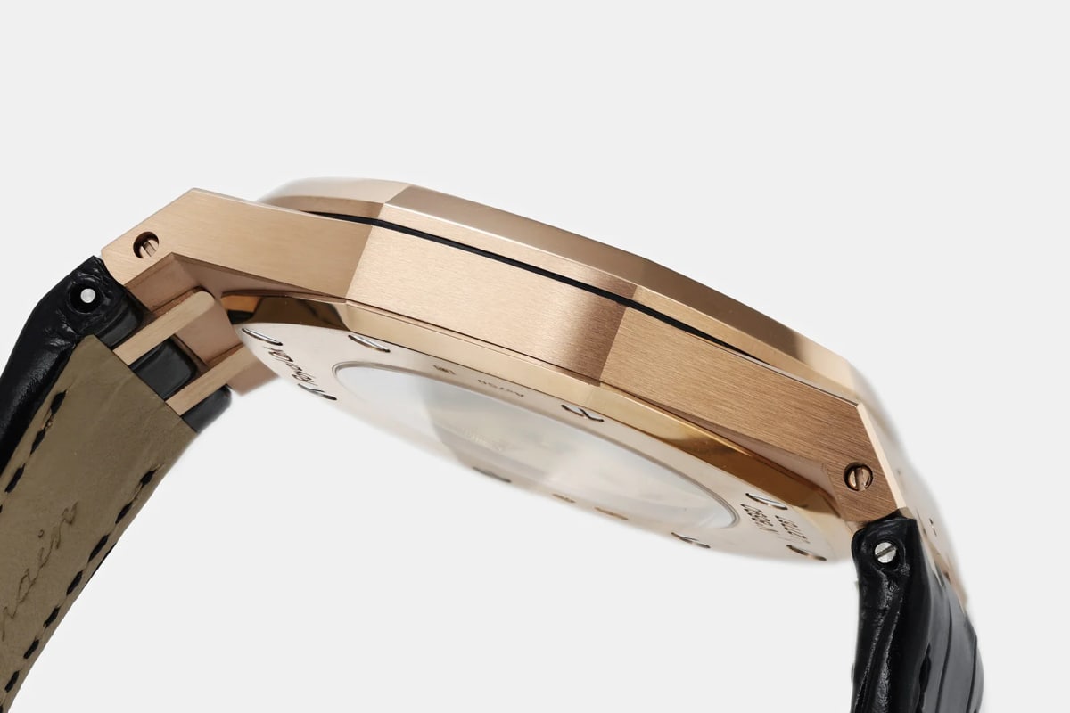 Case back and strap detail from below: the rose-gold case flank, the glass at the rear reflecting light, engraving around it, and the black leather strap with its pale lining and stitching