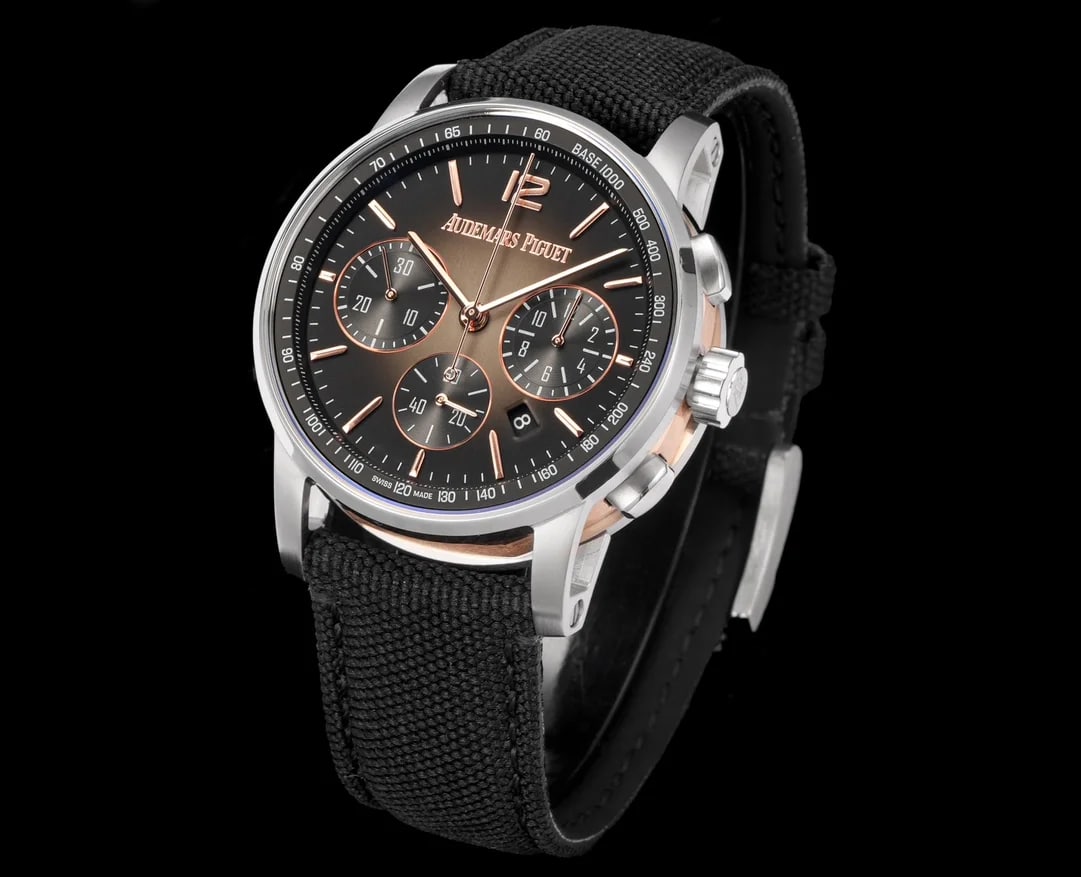Angled frame showing the round dial turned three-quarter so its sunburst fans out, the warm-toned mid case under a paler bezel, the crown and both pushers at the right and the black woven strap folded below