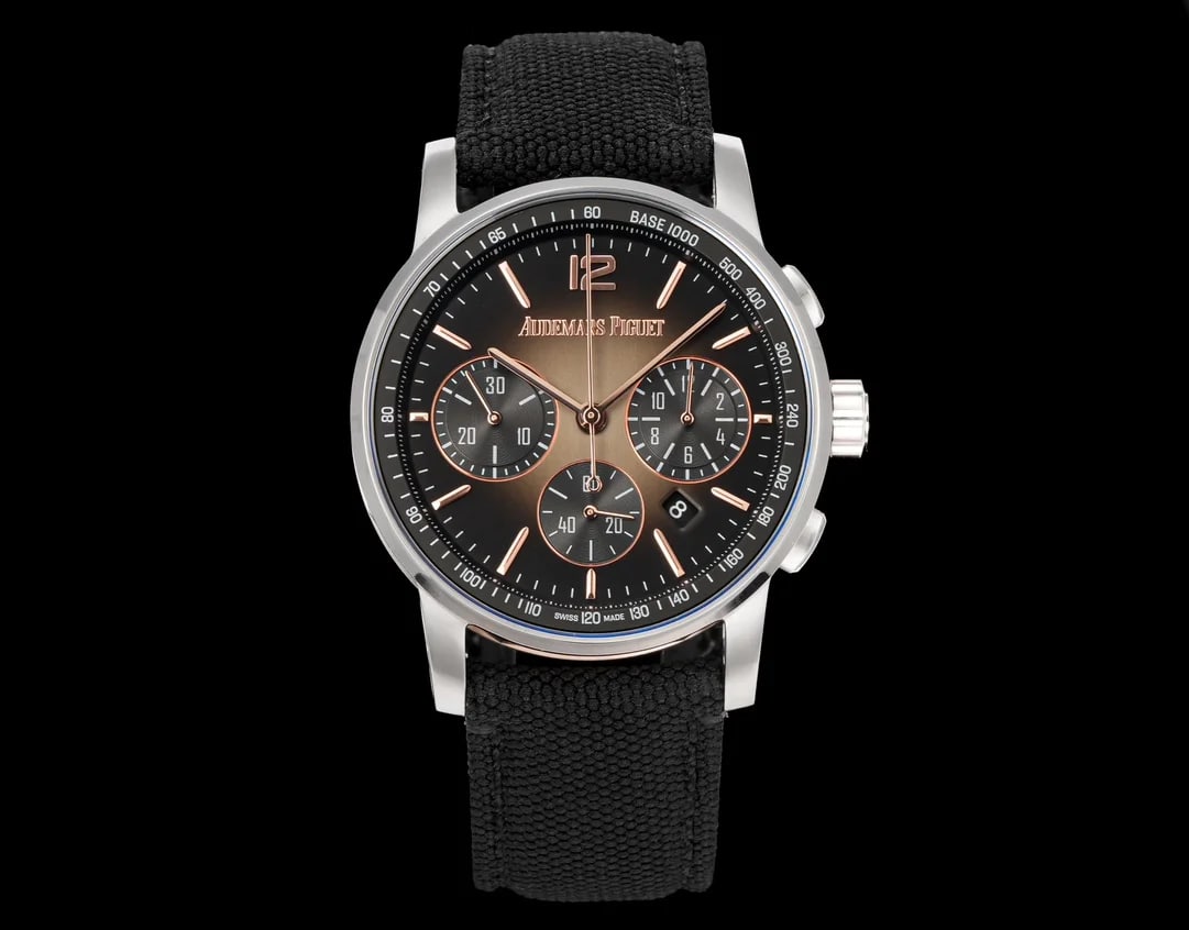 Smoked grey dial straight on inside a round case, darkening toward the edge, with three counters, the applied numeral twelve, a date aperture to the right and a black woven strap with pale stitching