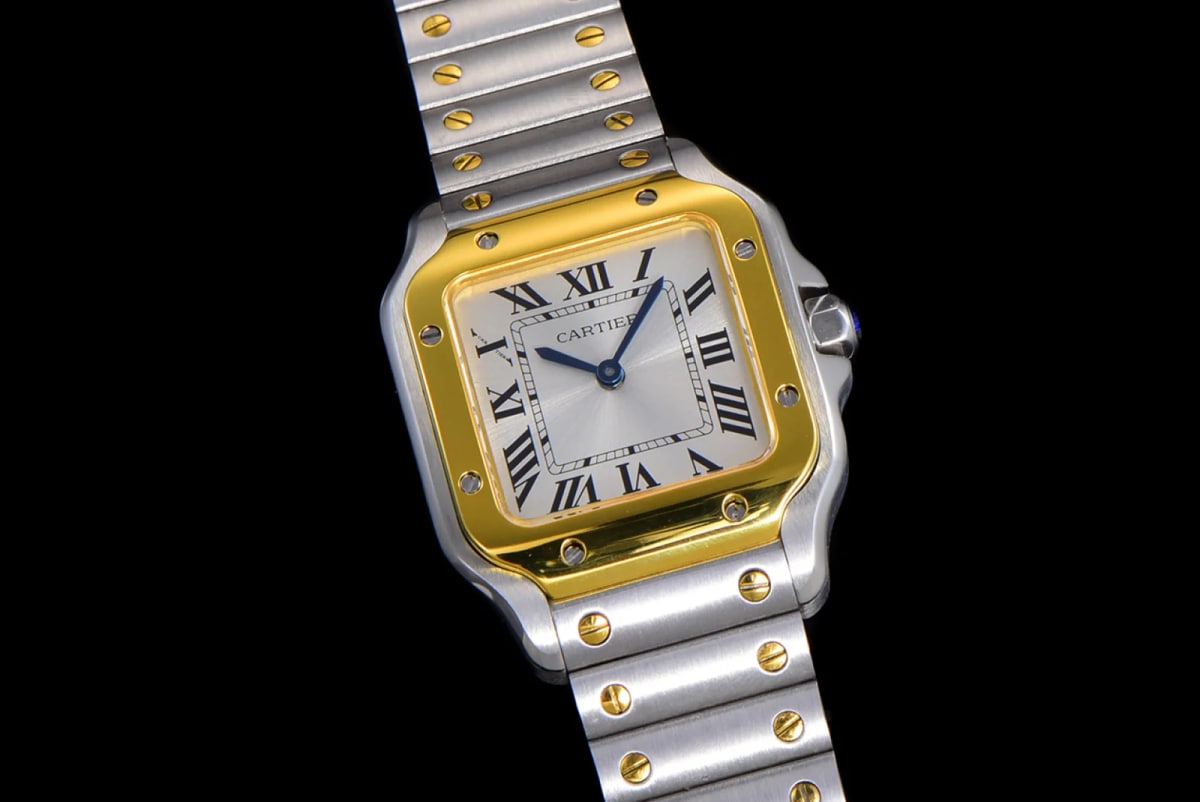 Angled three-quarter view from below, with the fluted crown and its blue cabochon at the right, the gold-toned bezel standing proud of the bowed case flank and the bracelet turning out of frame