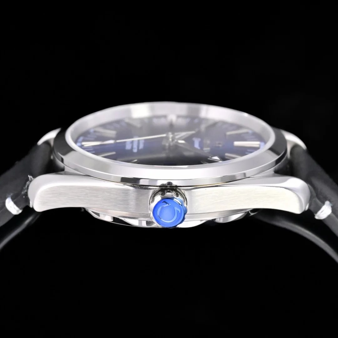 Case side detail with the crown standing in the foreground, its rounded blue centre under the light, the brushed case flank beside it, the blue dial seen almost edge-on behind the crystal, and the black textured strap running out of frame on both sides.