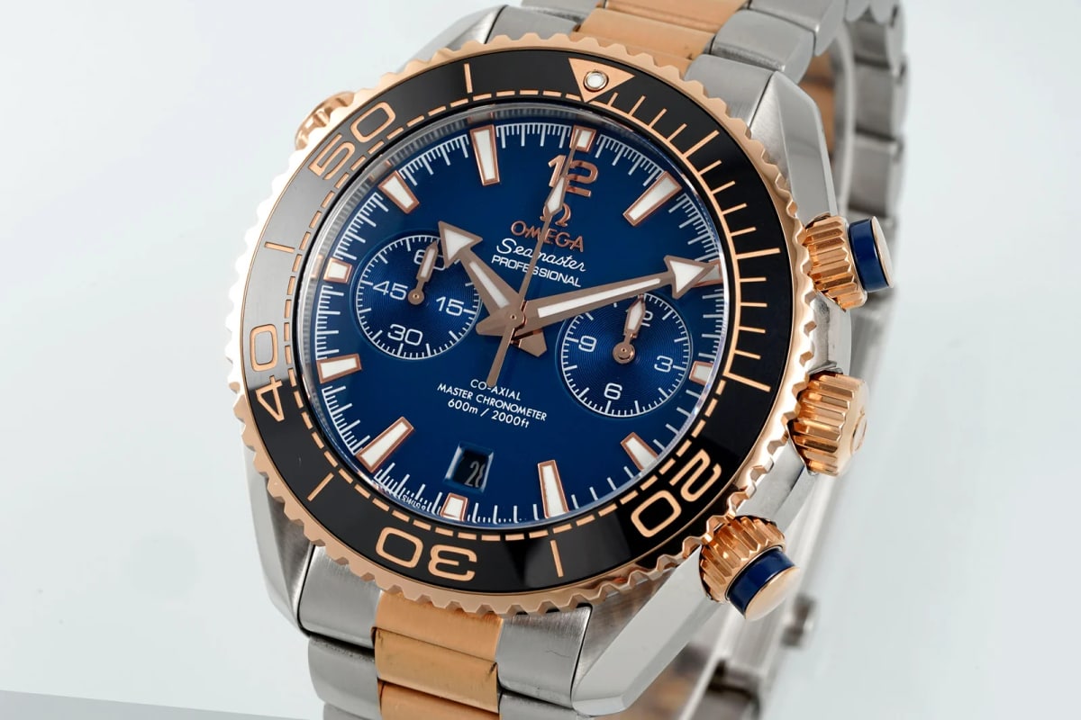 Angled three-quarter view with the bezel rim, crown and pushers standing at the right, the blue dial darkening toward the edge and the two-tone bracelet running down from the lugs