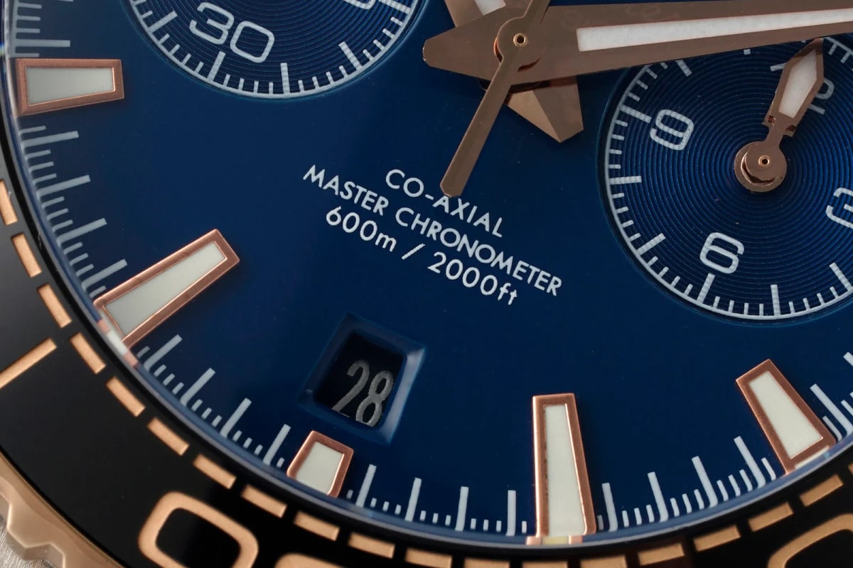 Dial close-up with the white CO-AXIAL MASTER CHRONOMETER 600m / 2000ft lettering above the date aperture showing 28, the gold-toned marker frames and the printed scale inside the right counter