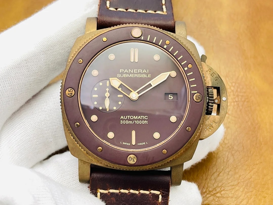 Brown dial straight on inside a bronze-toned cushion case held in a gloved hand, with luminous dot markers, printed numerals, the date at three, running seconds at nine and a dark brown stitched strap