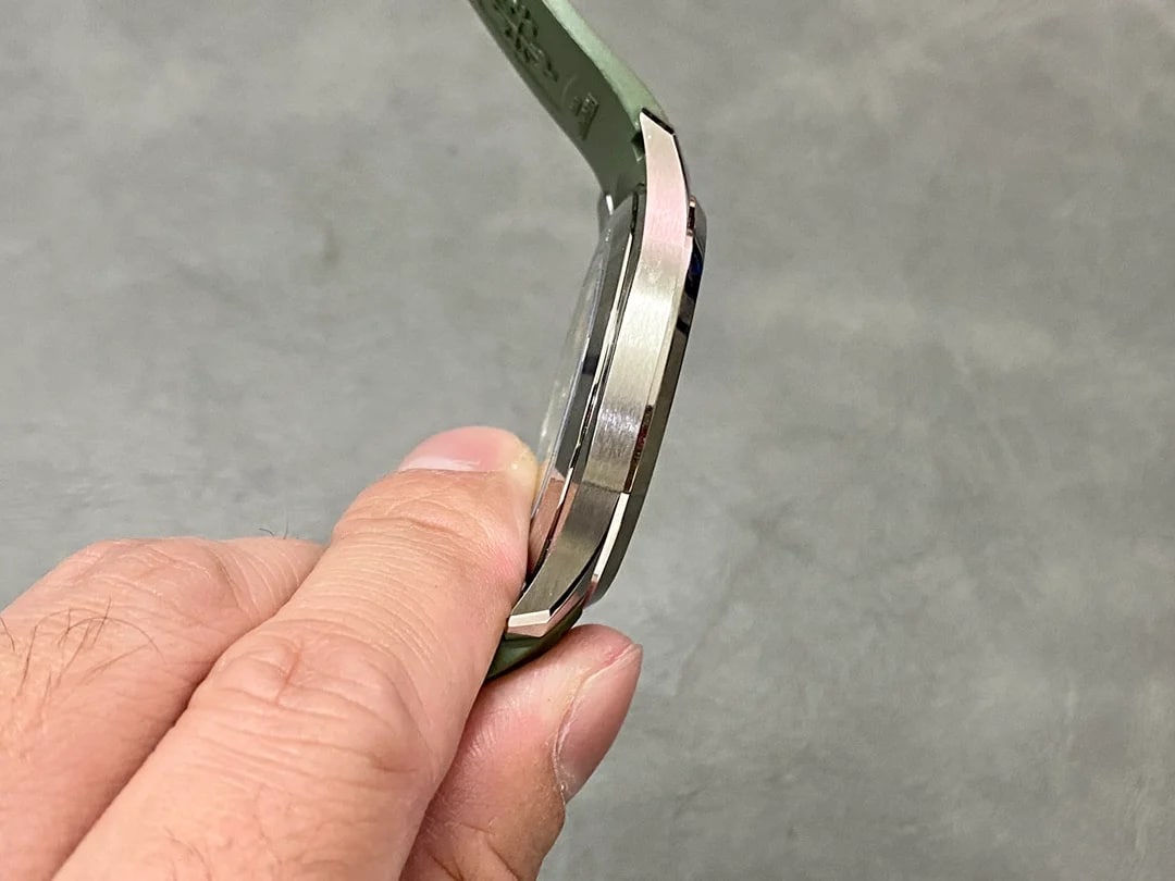 Close-up side view of the case held between the fingers: the case edge and its stepped flank, the crown on the right, and the green strap leaving the lugs with its raised grid pattern catching the light