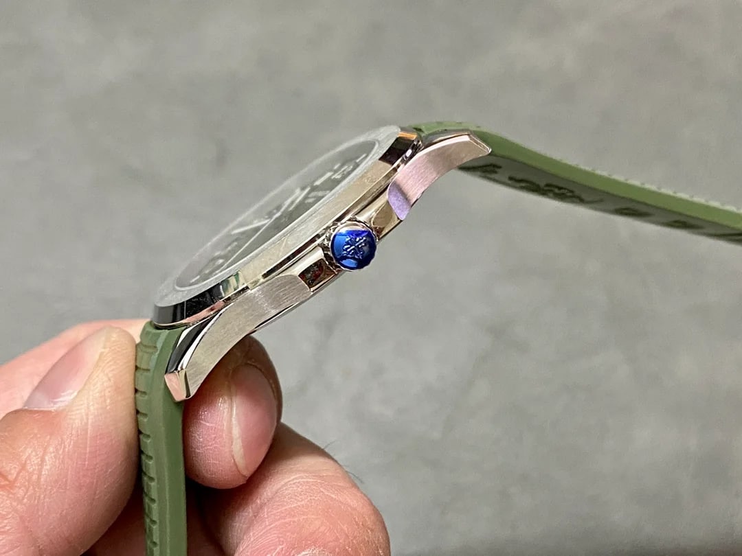 Crown detail seen along the case side: the winding crown with a blue stone standing proud of the flank, the stepped edge of the bezel above it, and the green strap with its raised grid pattern running out of frame