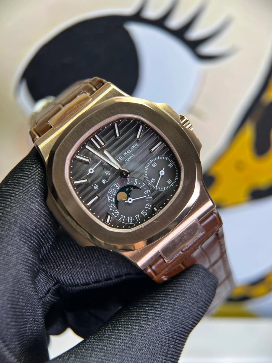 Three-quarter view of the case and brown dial, crown at the right, the moon-phase aperture low and left of centre, the date hand over printed numerals, the brown strap leaving the lugs