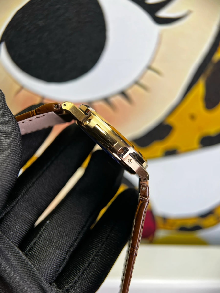 Clasp detail in profile: the thin gold-toned case edge, the crown on the flank, the folding clasp closed under a clear protective film and the brown strap with visible stitching