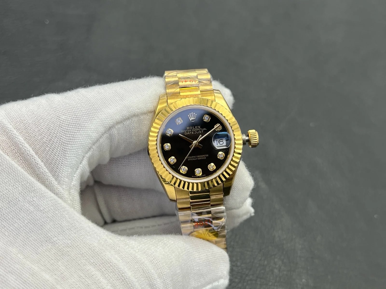 Black dial with stone markers facing the camera inside the fluted gold bezel, with the date under its magnifier and the three-piece bracelet below