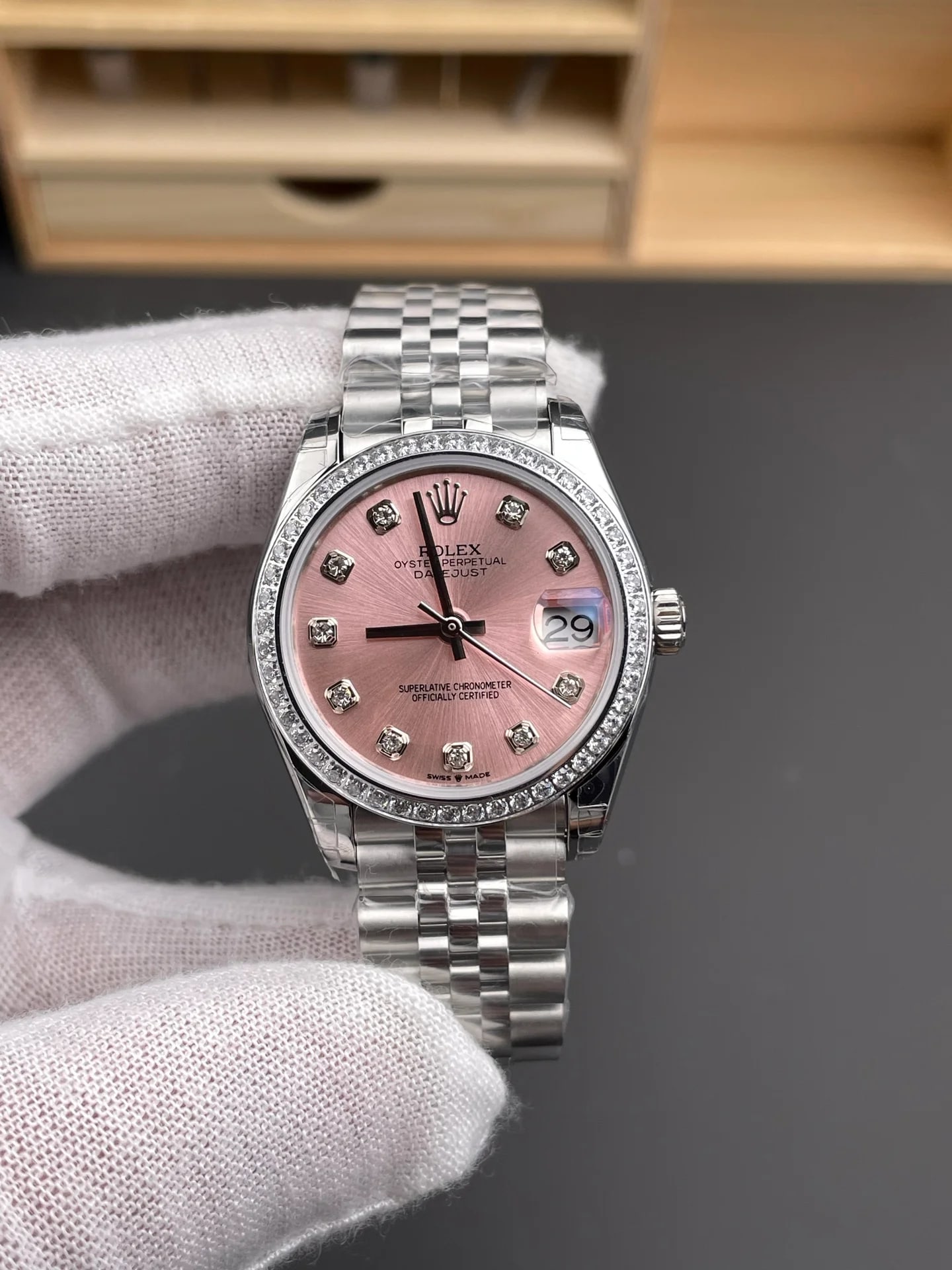 Three-quarter view of the stone-set bezel and the pink dial held in a gloved hand, with the five-link bracelet hanging below the case